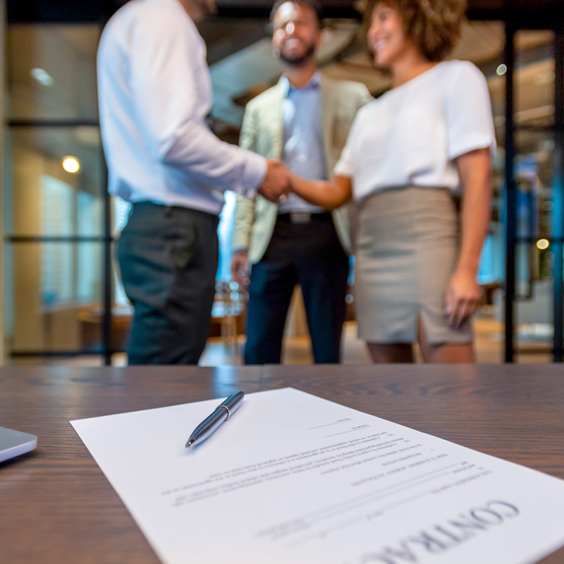 close up of contract sitting on table while people shake hands in the background